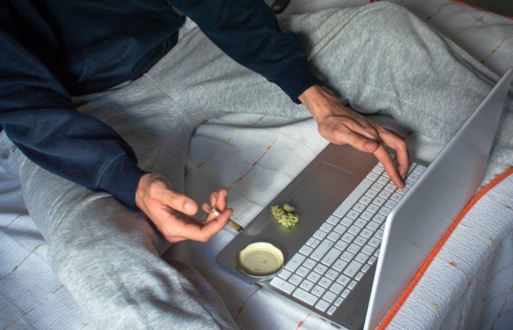 Person on a bed smoking marijuana joint while using a laptop to buy at an online dispensary in Canada. Speed Greens
